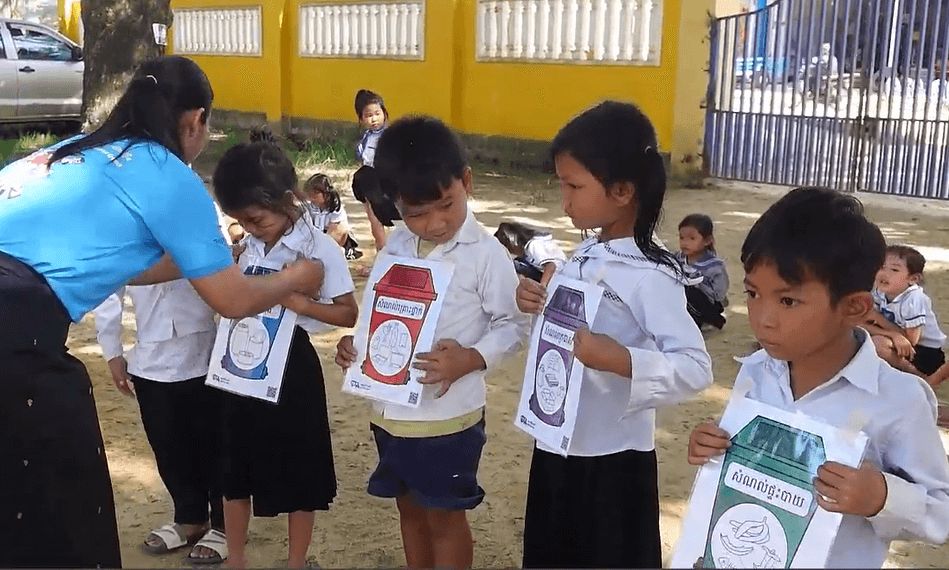 Children hold paper recycling-themed cards while a teacher guides an outdoor activity at their school