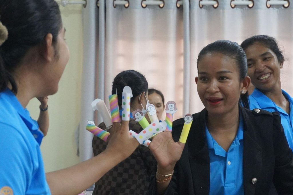 Teachers laugh while trying paper finger puppets on their fingers during an activity at the sixth workshop