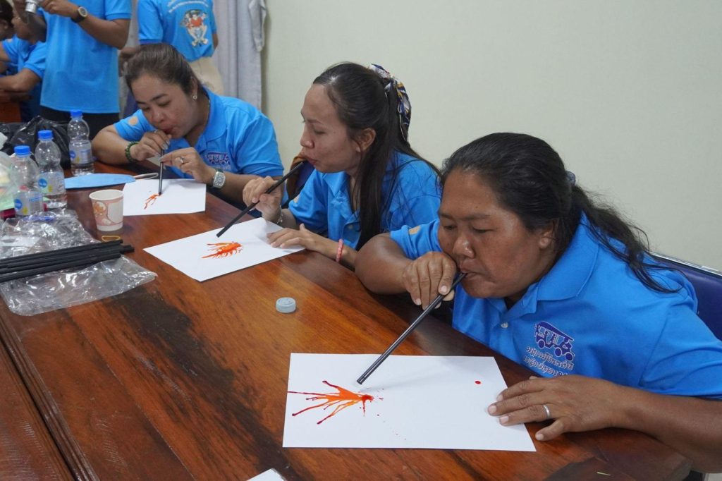 Three teachers blow paint through straws onto paper during a hands-on art activity at a workshop