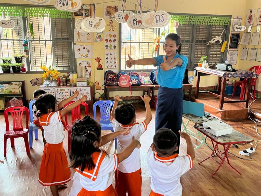 Children in orange and white uniforms follow their teacher’s arm movements in an action song