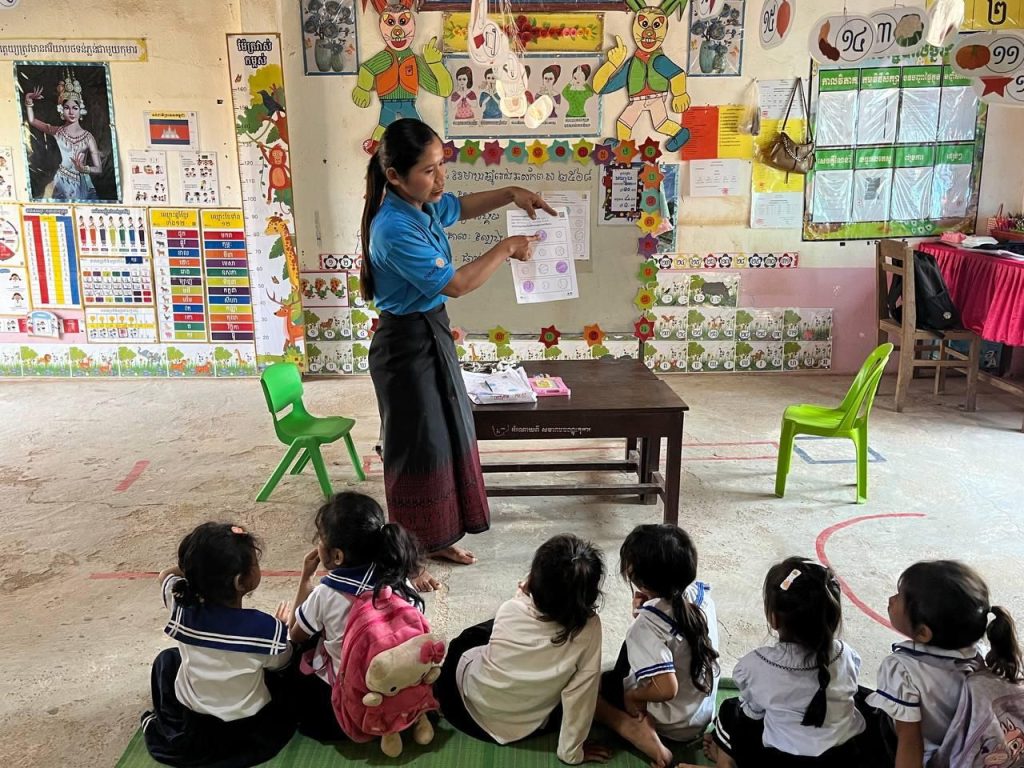 A teacher points to a picture card while preschool children sit on a mat in front of her