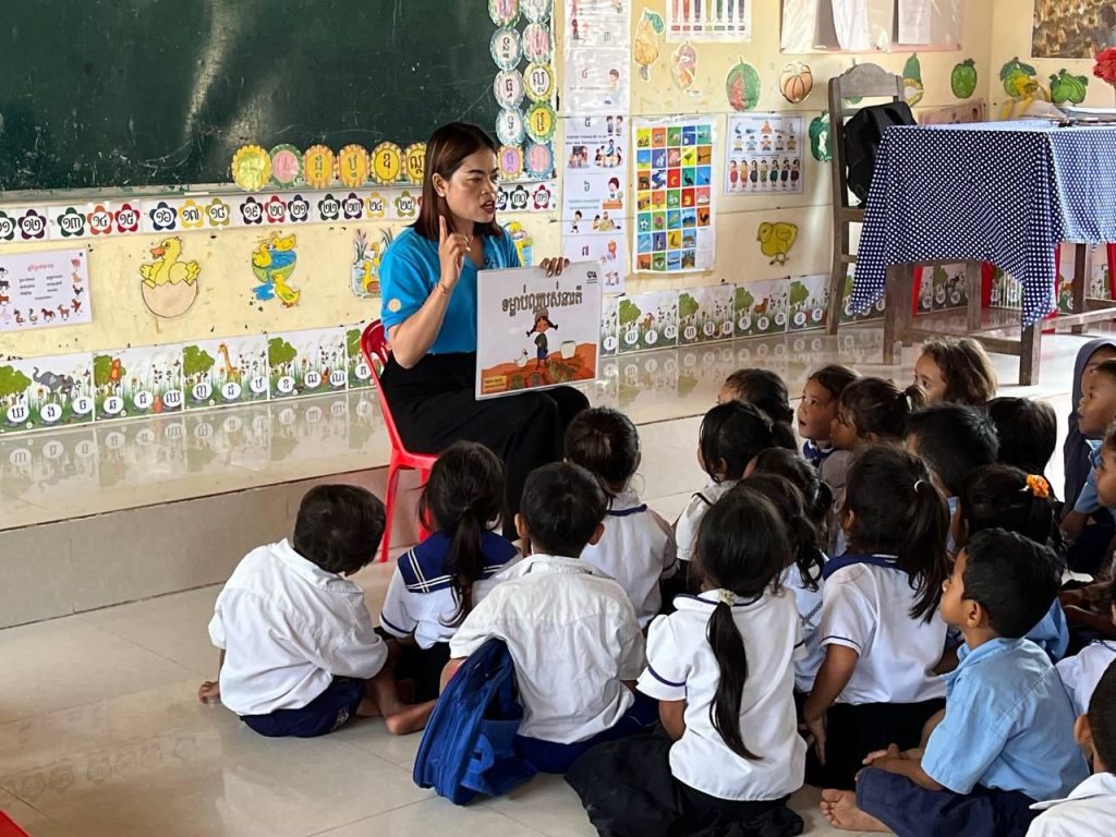 A teacher shows a picture book to children seated on the floor of a decorated classroom