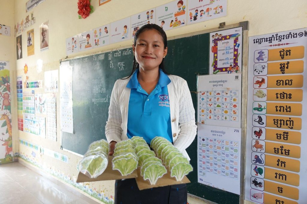 A teacher shows a tray of green paper crafts in front of a classroom blackboard