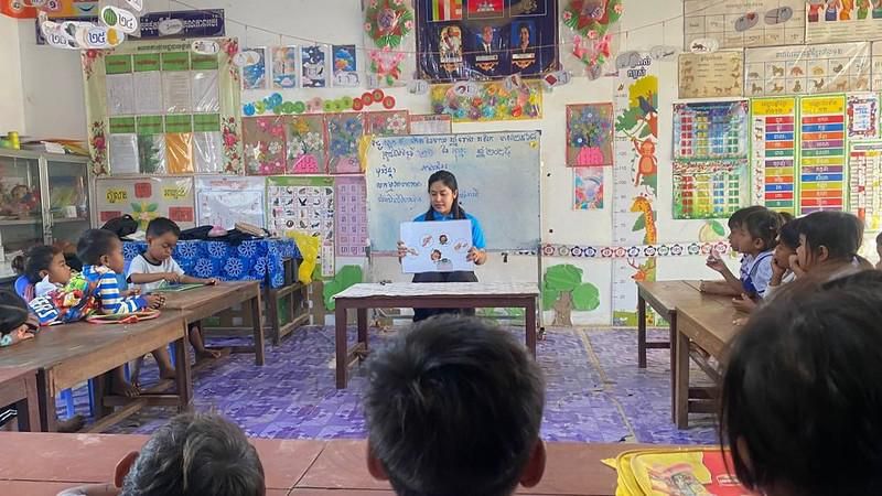 A teacher reads a picture book to a seated class from a table at the front of the room