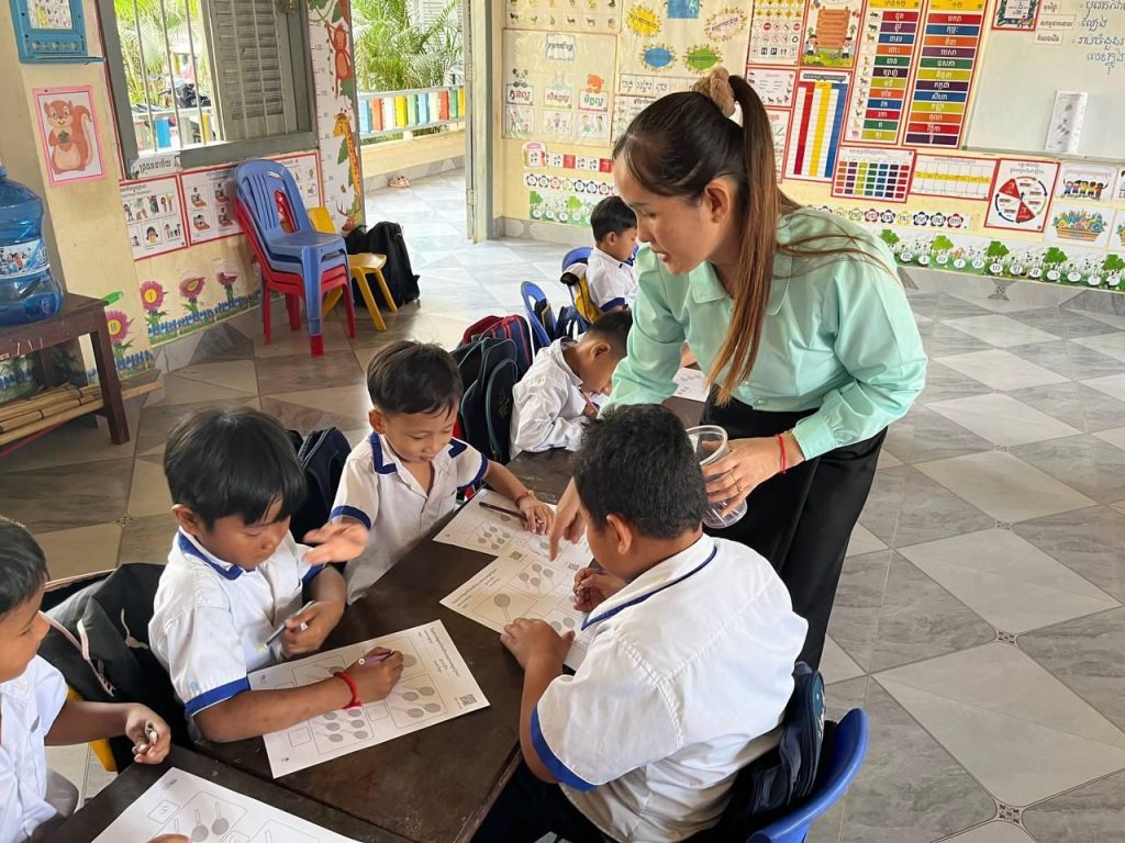 A teacher helps preschool children complete a worksheet at a table in a tiled classroom