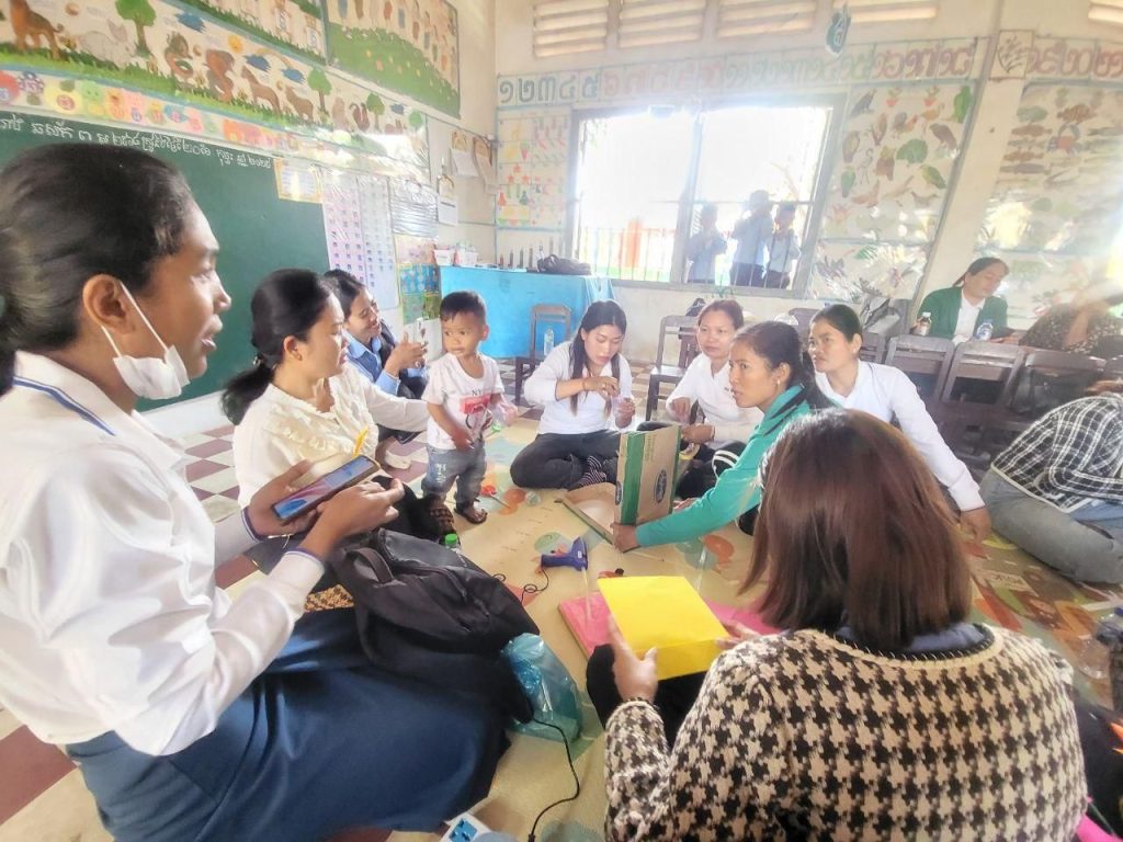 Teachers sit on the floor of a classroom sharing craft ideas during a technical meeting