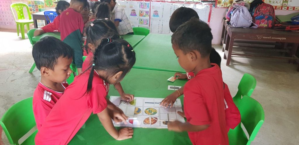 Children gather around a picture activity sheet on a green table in a classroom