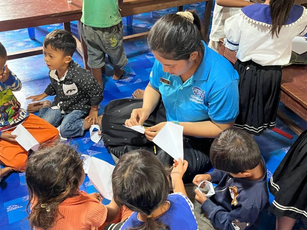 A teacher folds paper with children sitting around her on a blue mat