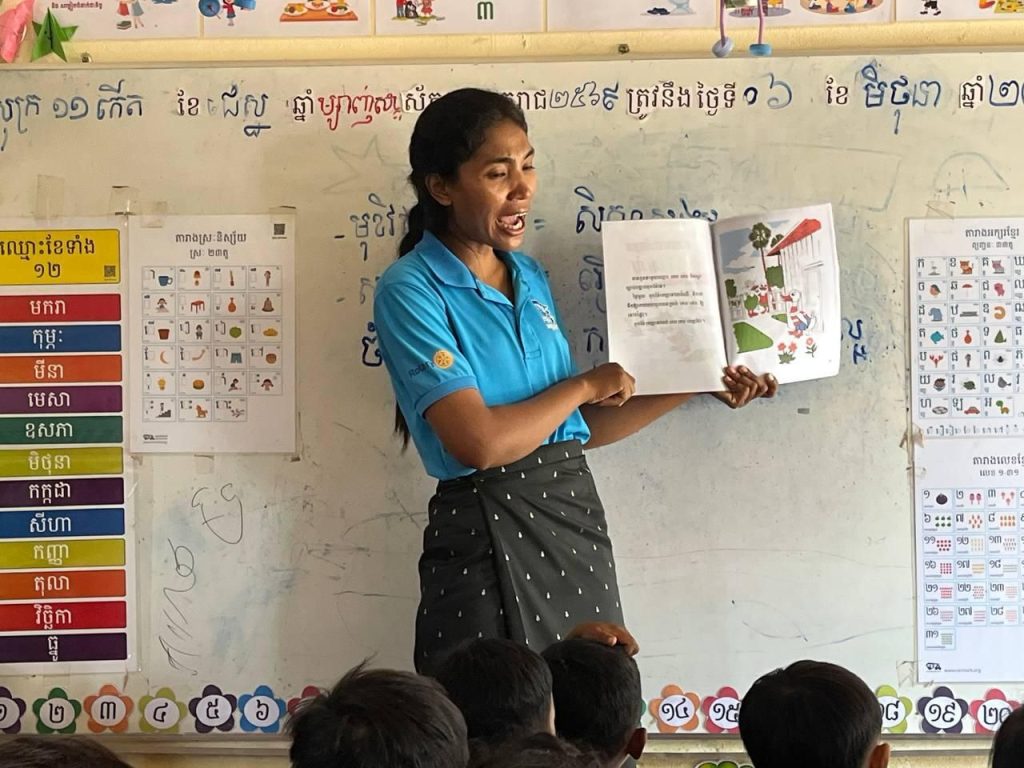 A teacher shows pages of a picture book while standing in front of a whiteboard