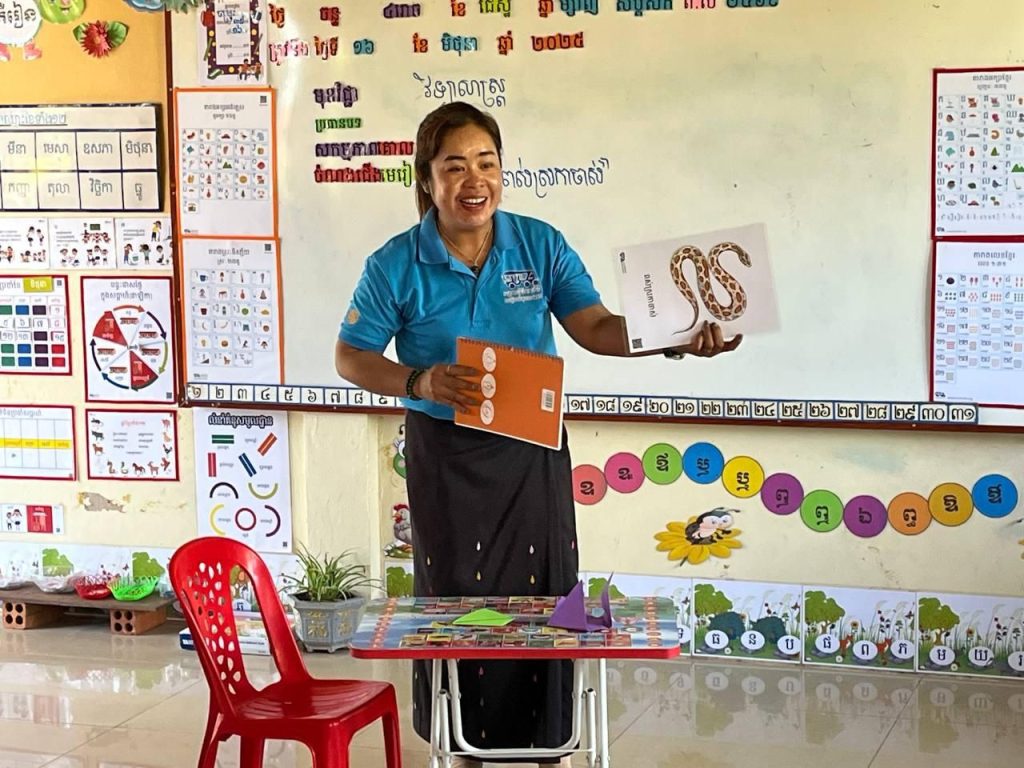 A teacher holds up a snake picture card and a wooden card at the front of her classroom