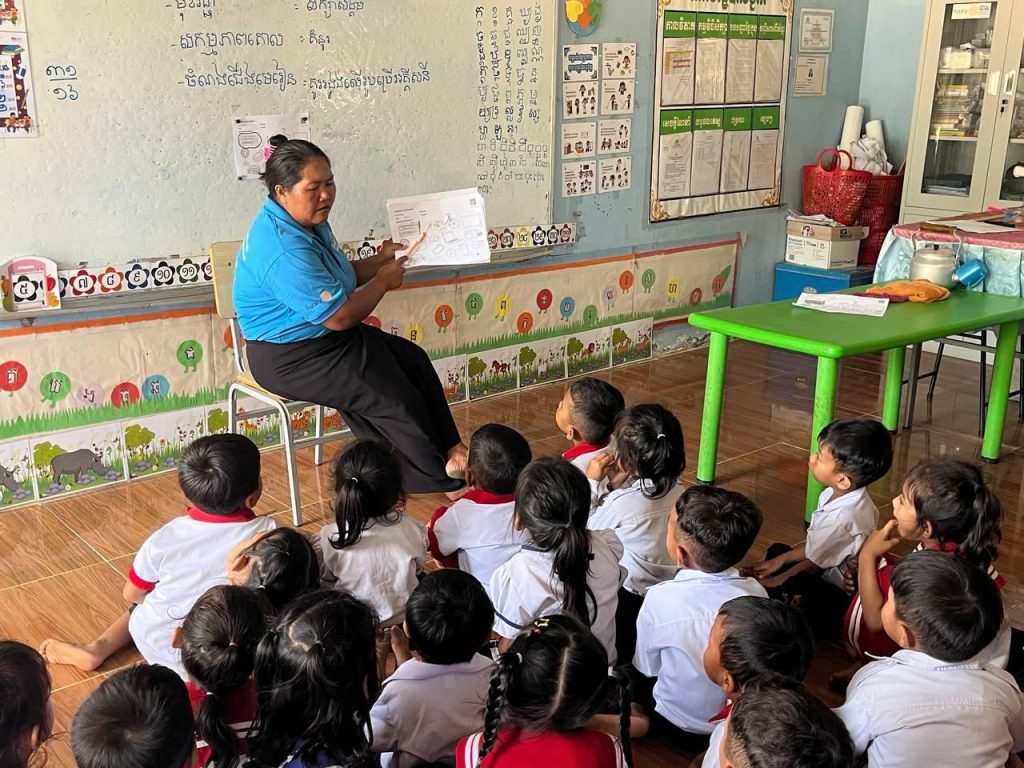 A teacher reads a worksheet to children seated on the floor of a brightly decorated classroom