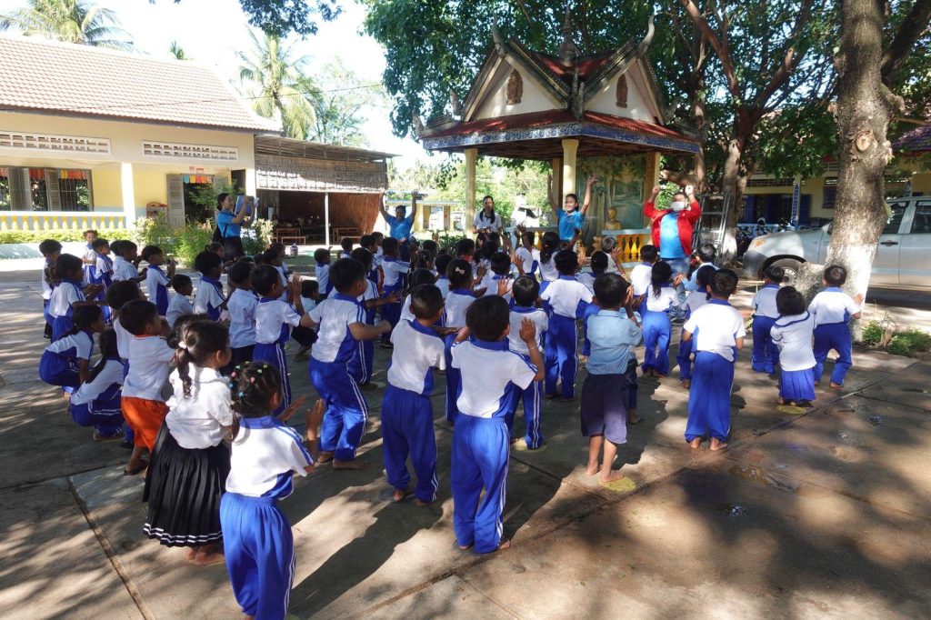 Children in school uniforms gather in a shaded school yard for an outdoor activity