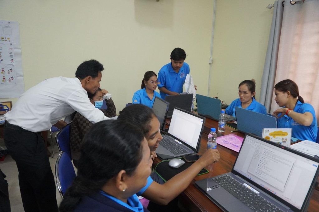 A trainer leans over to help teachers at laptops while others work on worksheets