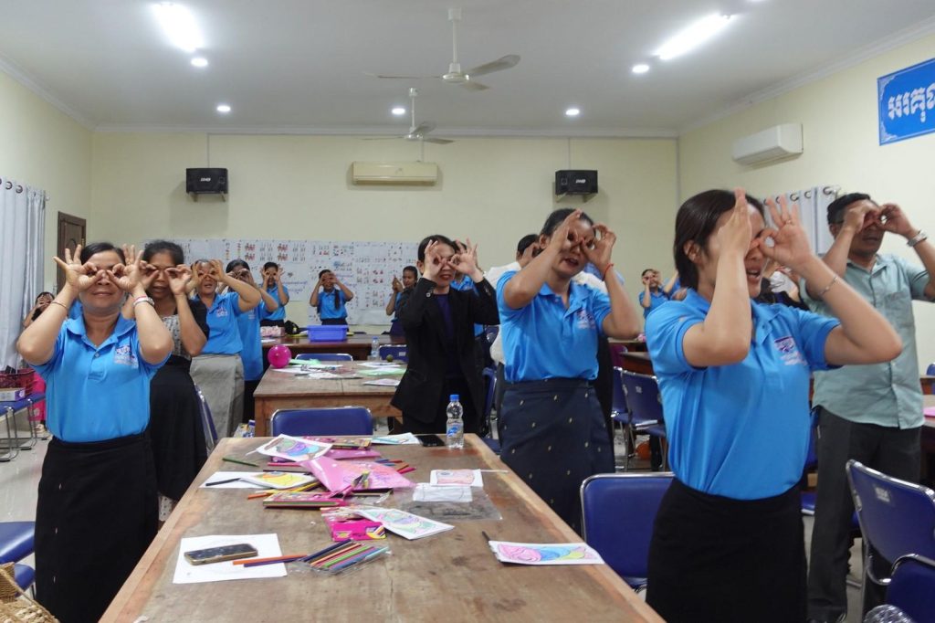 Teachers copy a hand-play song movement with hands around their eyes in the workshop room