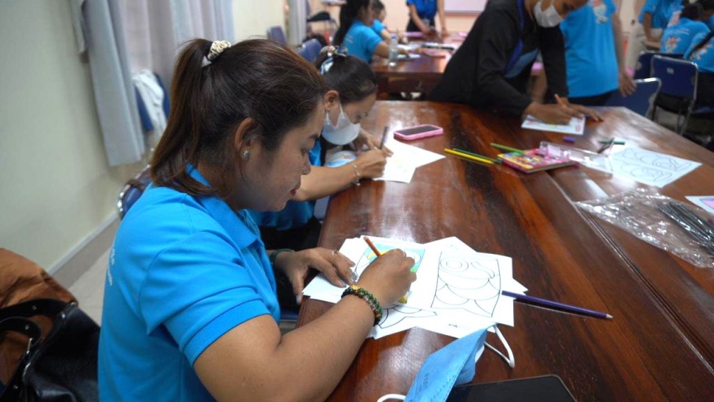 A teacher colors in a worksheet with a pencil at a long table during the workshop