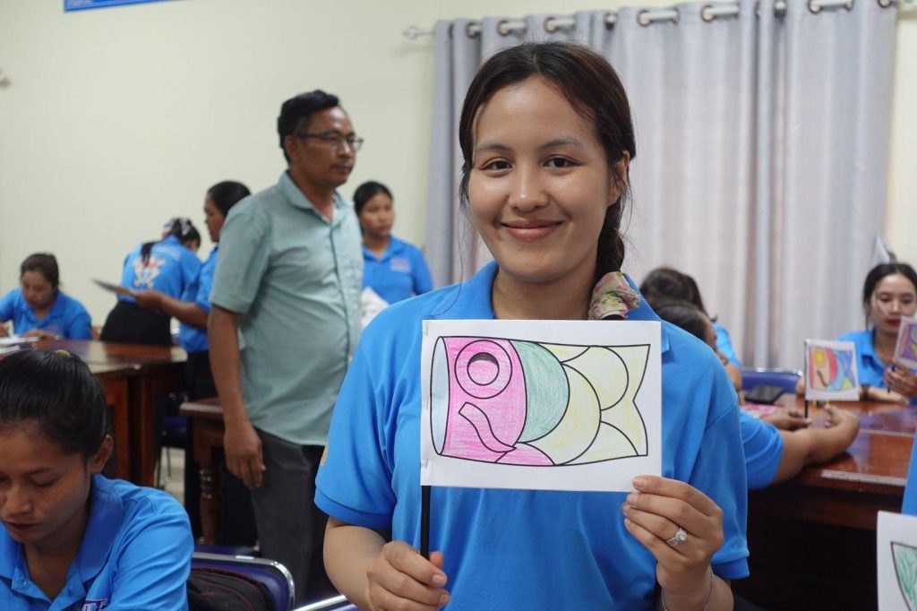 A teacher holds up her finished colored fish drawing while others work behind her