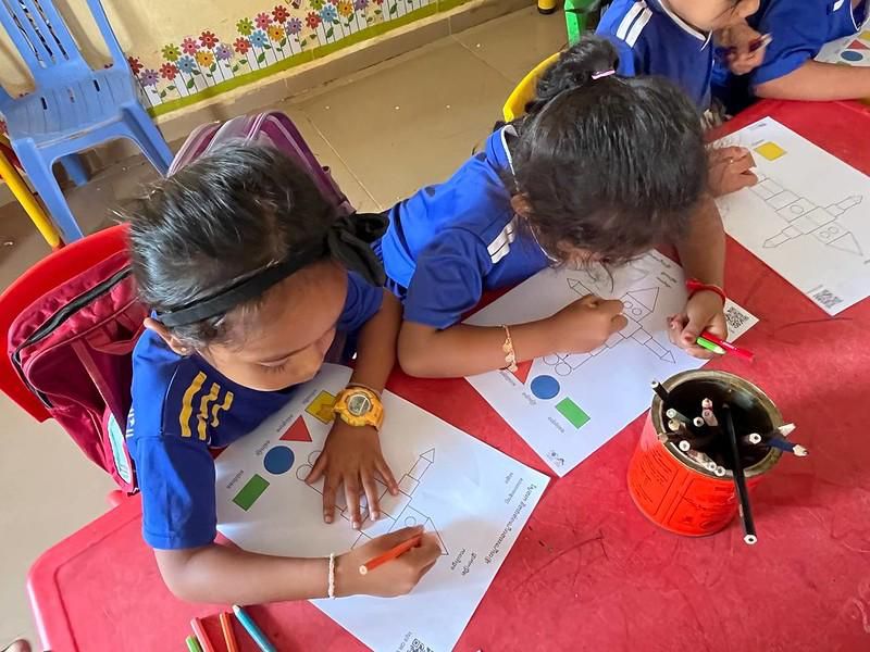Two children color shapes on worksheets at a red table in a classroom