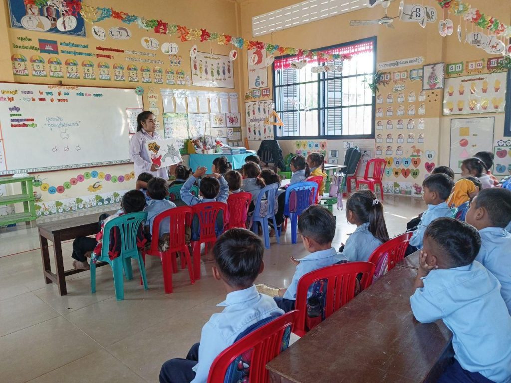 Children sit at tables in a bright classroom while a teacher teaches at the front