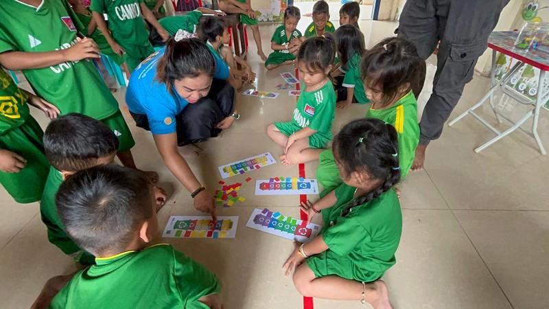 A teacher kneels with children in green uniforms playing a board game on the floor