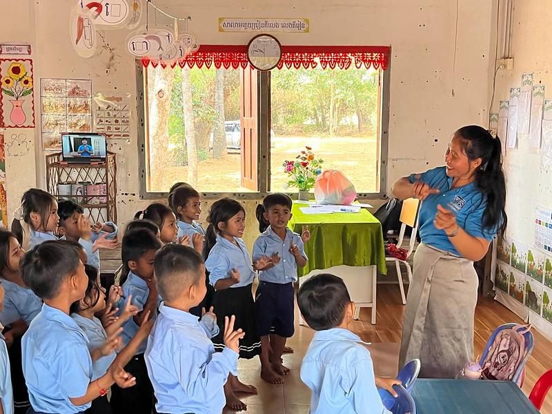 A teacher leads children in a hand-play song in front of a classroom screen