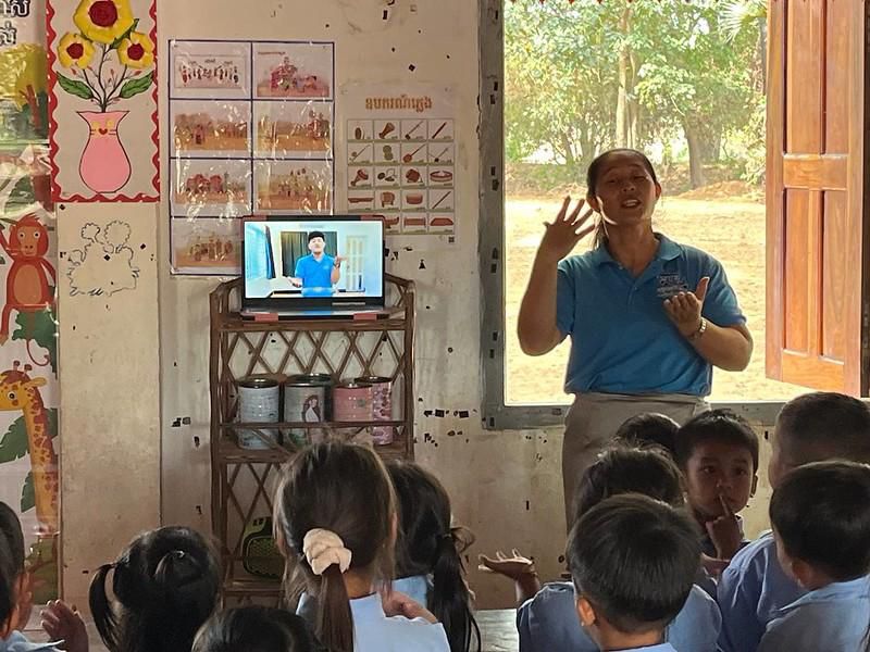 A teacher gestures while a video plays on a screen behind her in a classroom with seated children