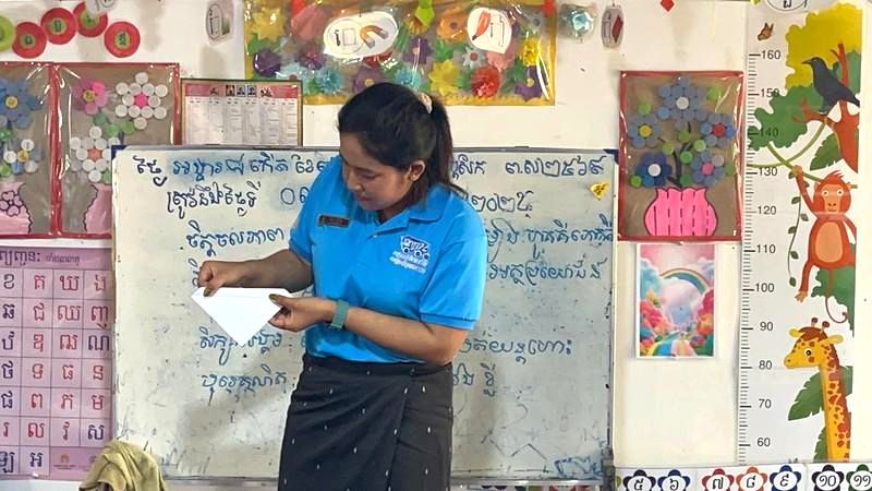 A teacher folds a paper craft in front of a blackboard covered in handwriting