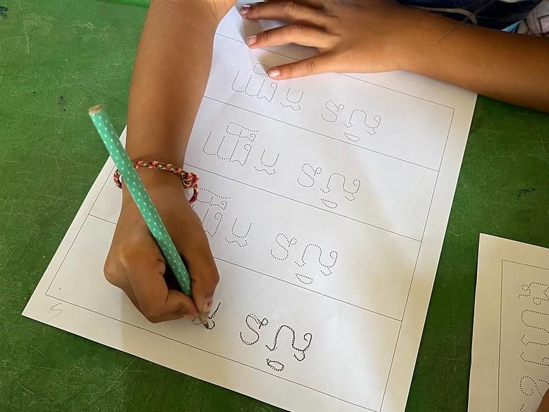 A child traces dotted Khmer letters on a worksheet with a green pencil