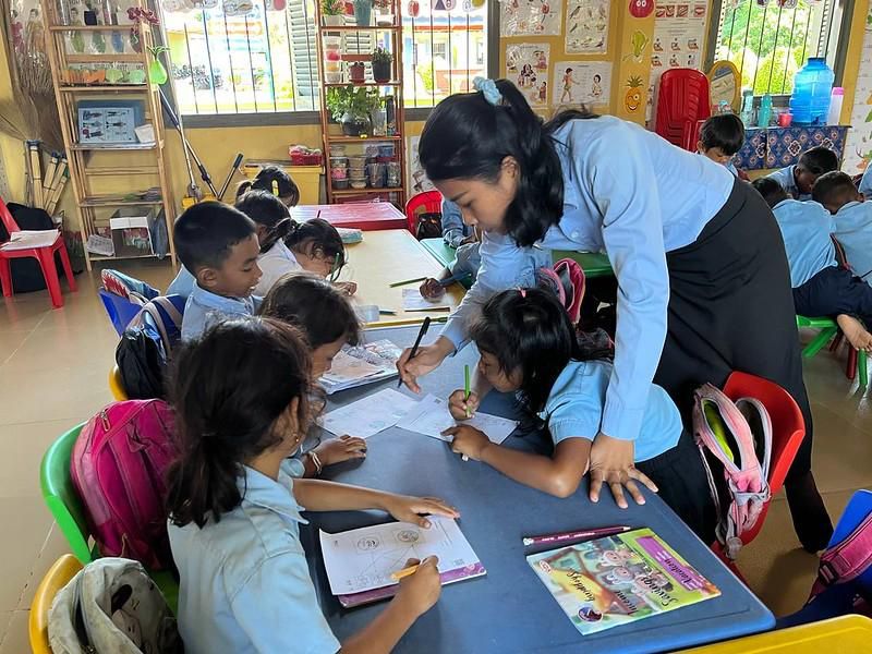 A teacher bends over children drawing at a blue table in a classroom during a school visit