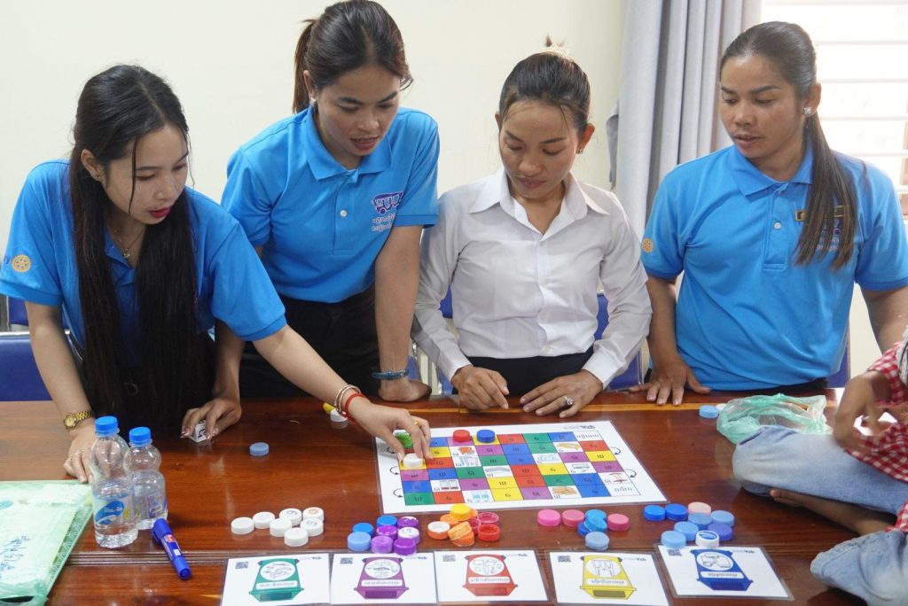 Teachers lean over a colorful board game with bottle caps on a wooden table