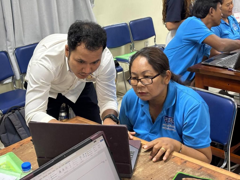 A trainer leans over to help a teacher at her laptop during the final computer session