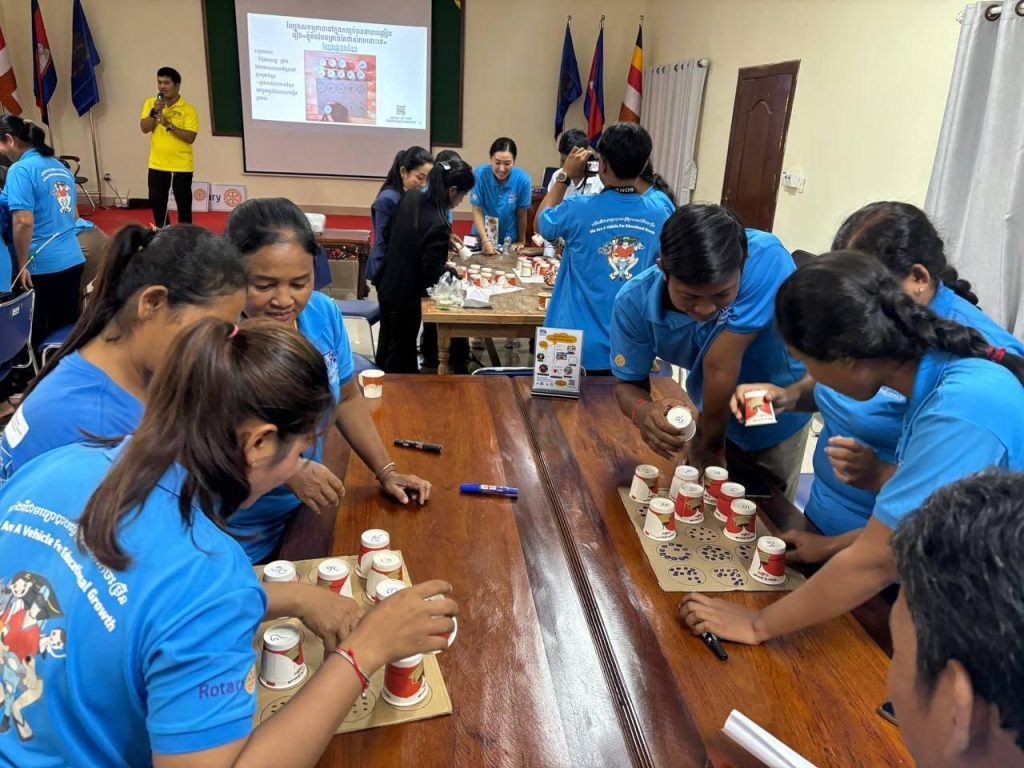 Teachers gather around a table making a cup game while a presentation shows on the screen