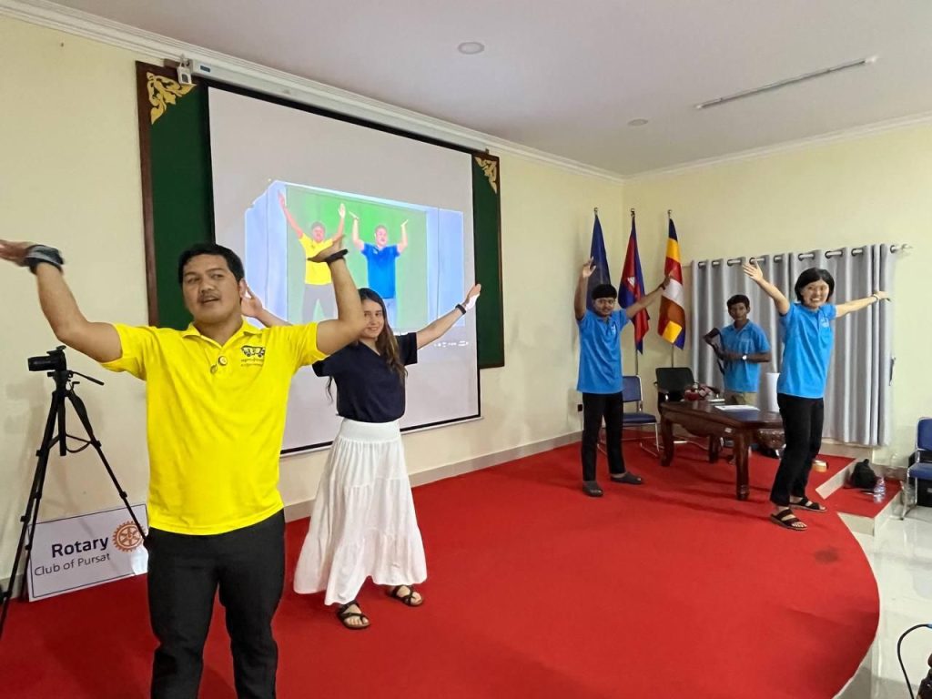 A man in a yellow shirt and others dance to a video shown on the screen in the workshop room