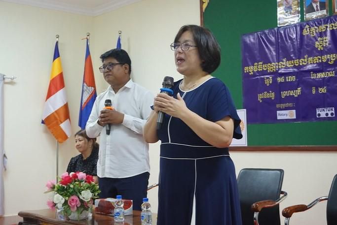 A Japanese expert in a navy dress speaks into a microphone beside a man at the front of the room