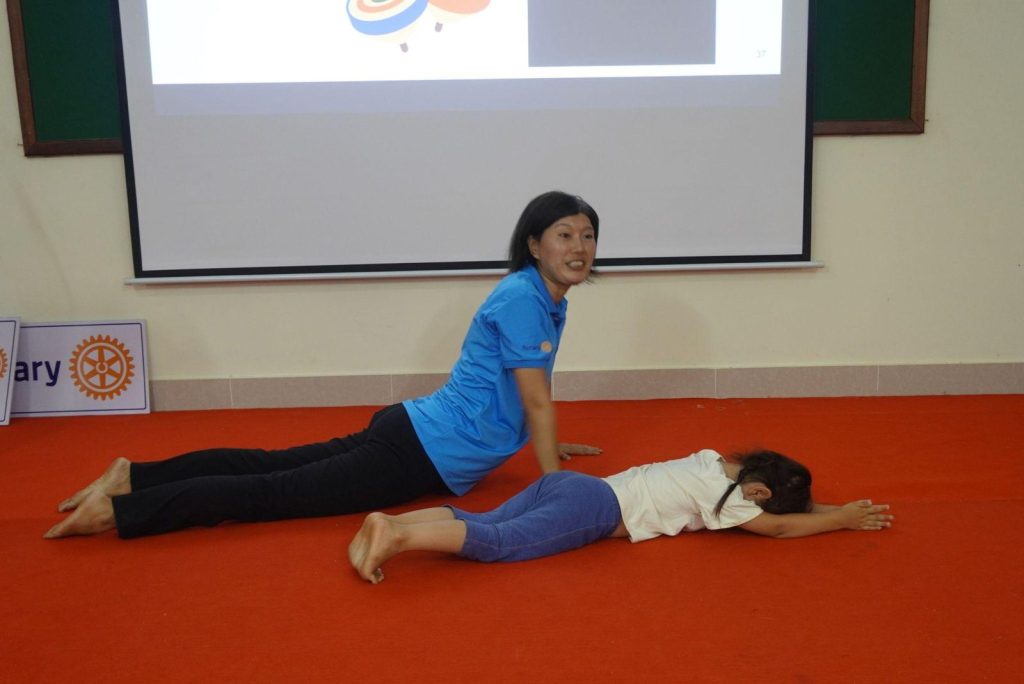Two people lie on the red floor in front of the screen while demonstrating a body exercise