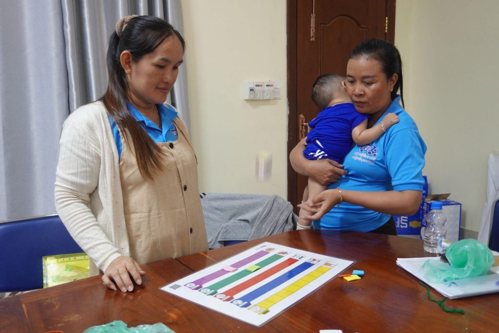 A teacher holding a small child looks at a striped paper game board on a table