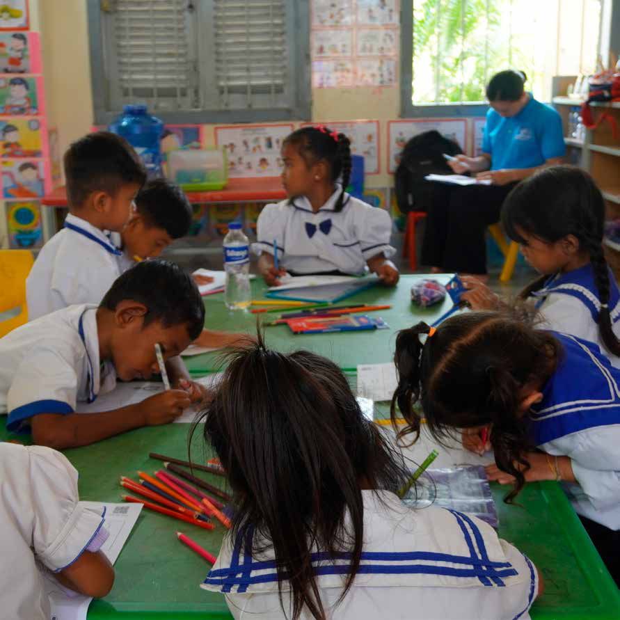 Children in school uniform sit around a green table drawing with coloured pencils in a classroom.