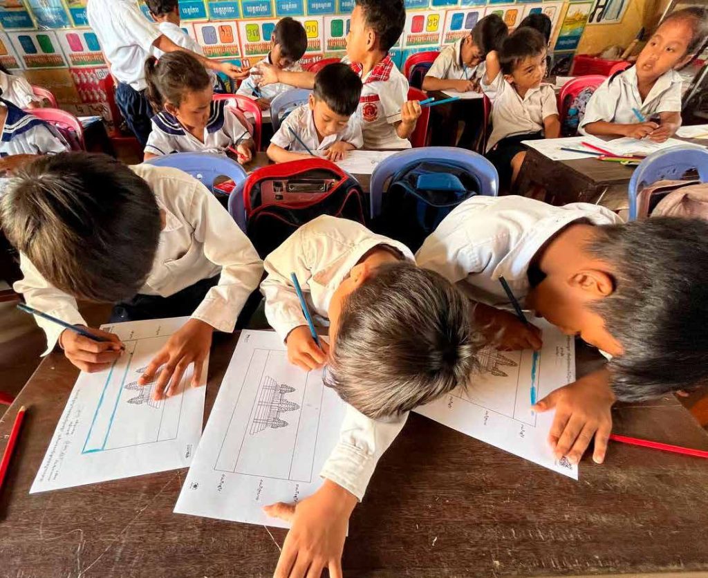 Children in school uniform bend over desks writing and drawing on worksheets in a crowded classroom.