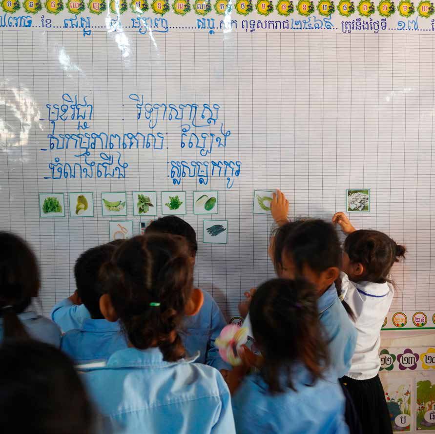 Young children stand at a classroom whiteboard covered in Khmer writing and small picture cards during a lesson.