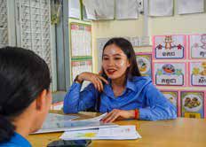A smiling teacher in a blue shirt sits at a desk in a classroom during one-on-one support.