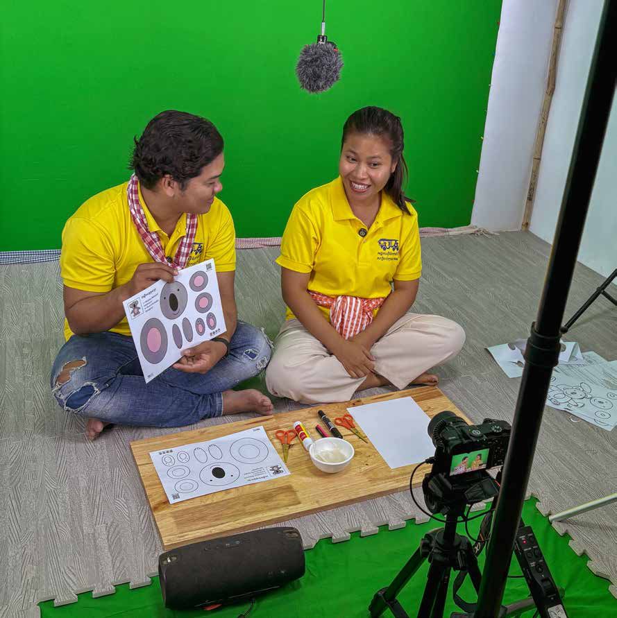 Two presenters in yellow shirts sit on the floor in front of a green screen, holding a craft for filming.