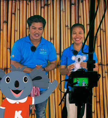 Two presenters in blue shirts smile in front of a bamboo wall with a camera and a koala character.