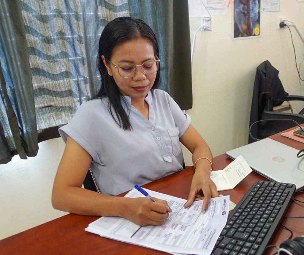 A TukTuk team member in a grey top writes on a form at her desk beside a keyboard.