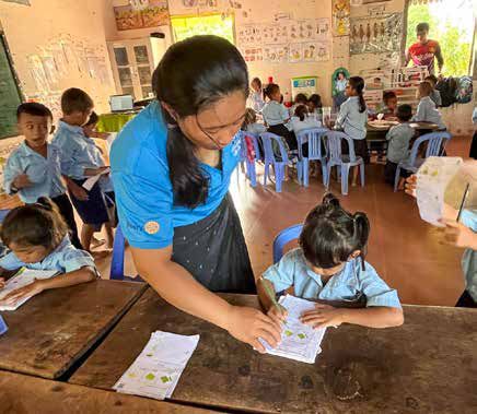 A teacher in a blue shirt bends over a desk to help a preschool child with a worksheet.