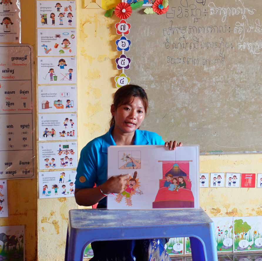 A teacher in a blue shirt holds up an illustrated story picture in a classroom with wall charts.