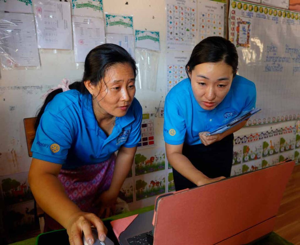 Two teachers in blue shirts lean over a laptop together in a classroom with learning charts on the wall.