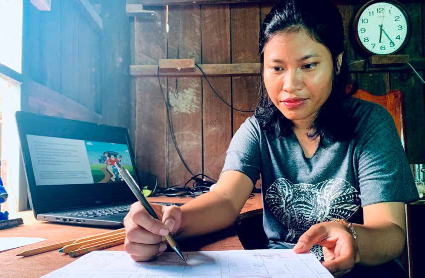 A team member sits at a wooden desk with a laptop and writes notes while creating learning resources.