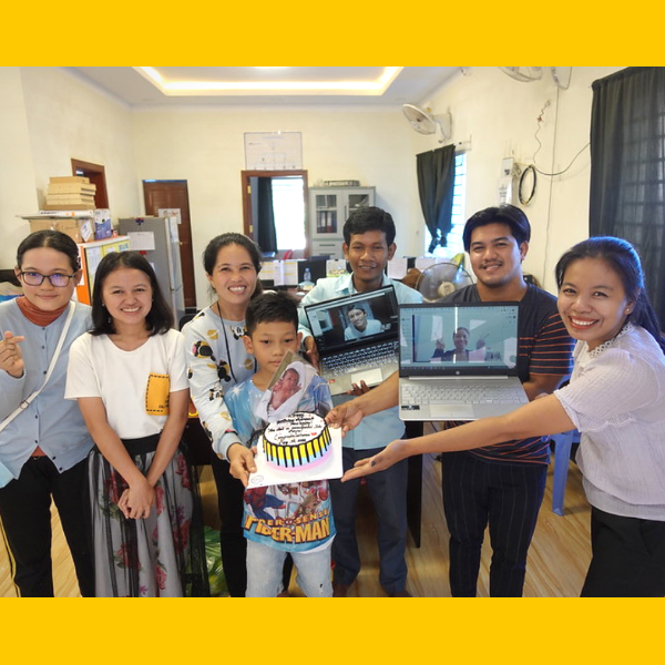 TukTuk staff gather in the office around a birthday cake while colleagues join the celebration by video call