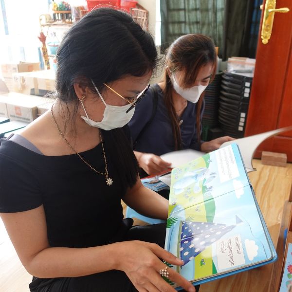 A teacher in a face mask looks through a TukTuk picture book while a colleague reads beside her