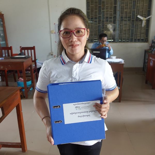 A woman holds up a blue folder of TukTuk teaching materials in a school office