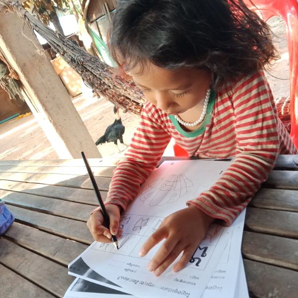 A girl in a striped top writes on a worksheet at an outdoor table while a chicken wanders behind her